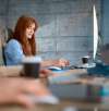 Smiling woman with red hair working at a desktop computer, taking notes in a bright office with coffee cups nearby.