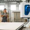 Worker in a cap and overalls using a tablet to monitor a machine in a furniture manufacturing facility