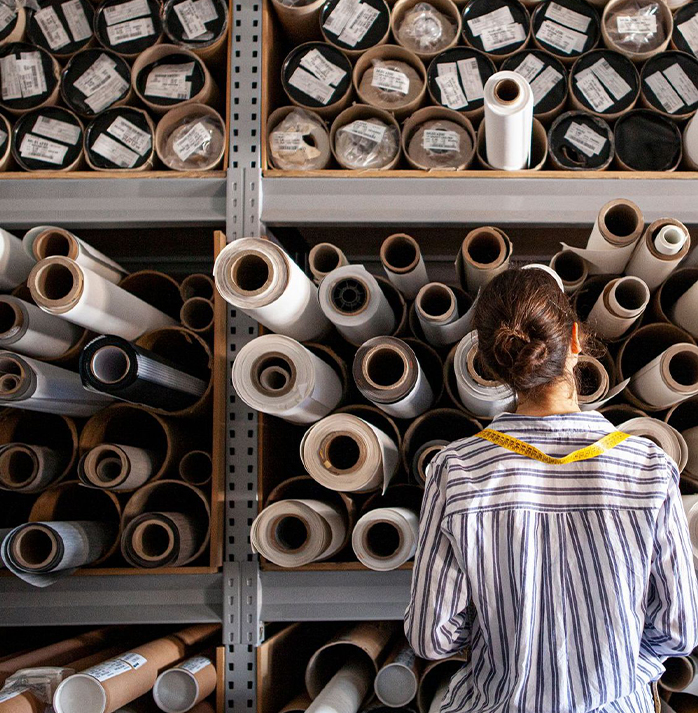Person with a measuring tape around their neck, standing in front of shelves filled with rolled fabrics.