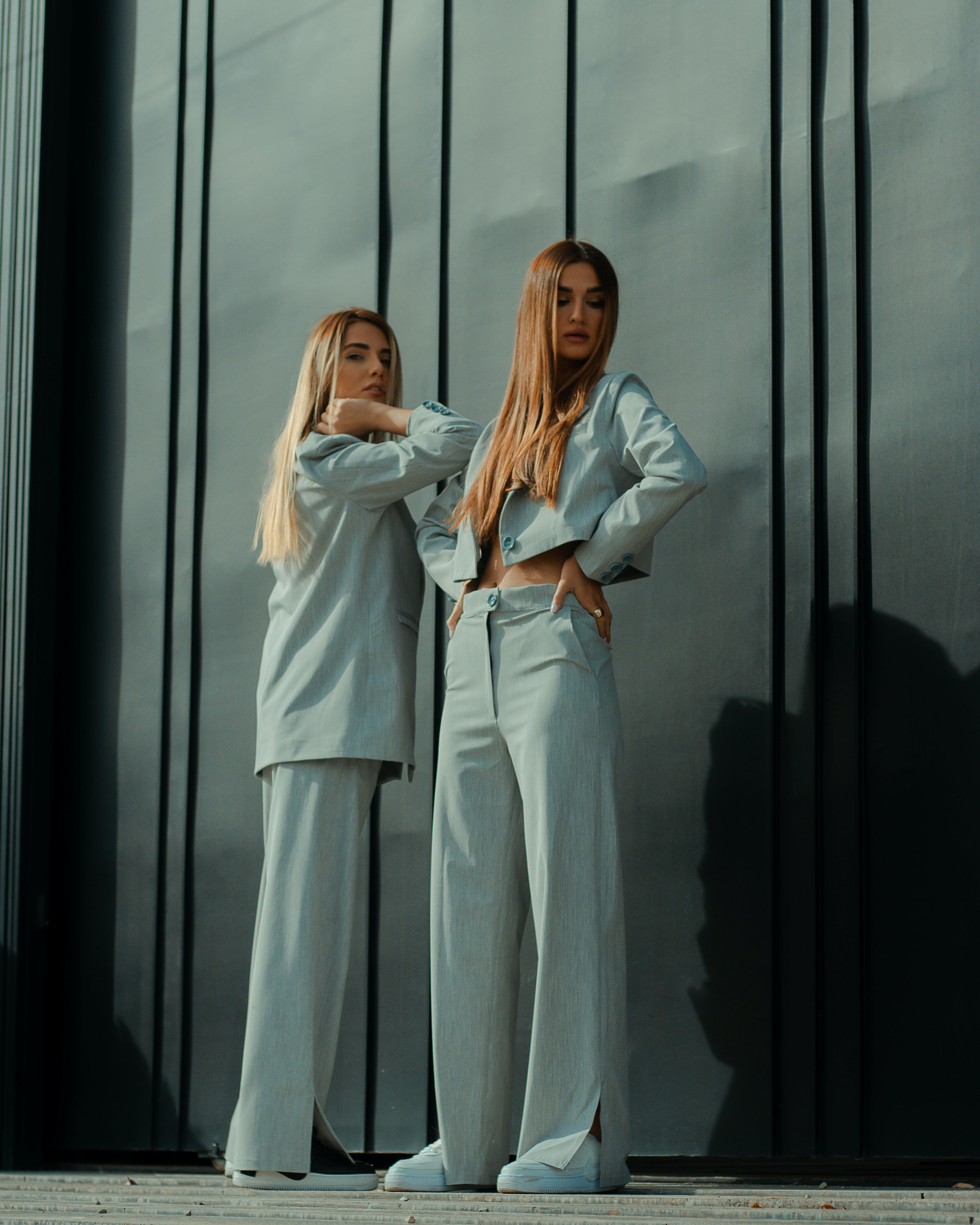 Two women in matching light gray tailored suits, standing confidently against a modern, dark vertical panel backdrop
